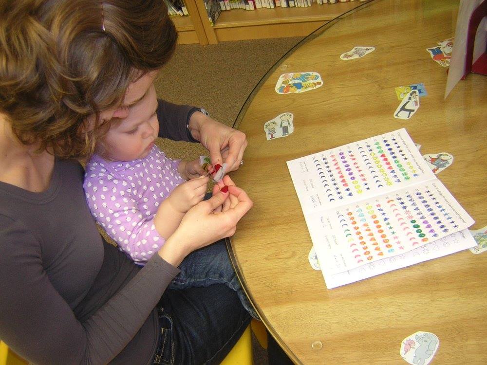 Mother helps toddler put stickers into the 1000 books before Kindergarten booklet.